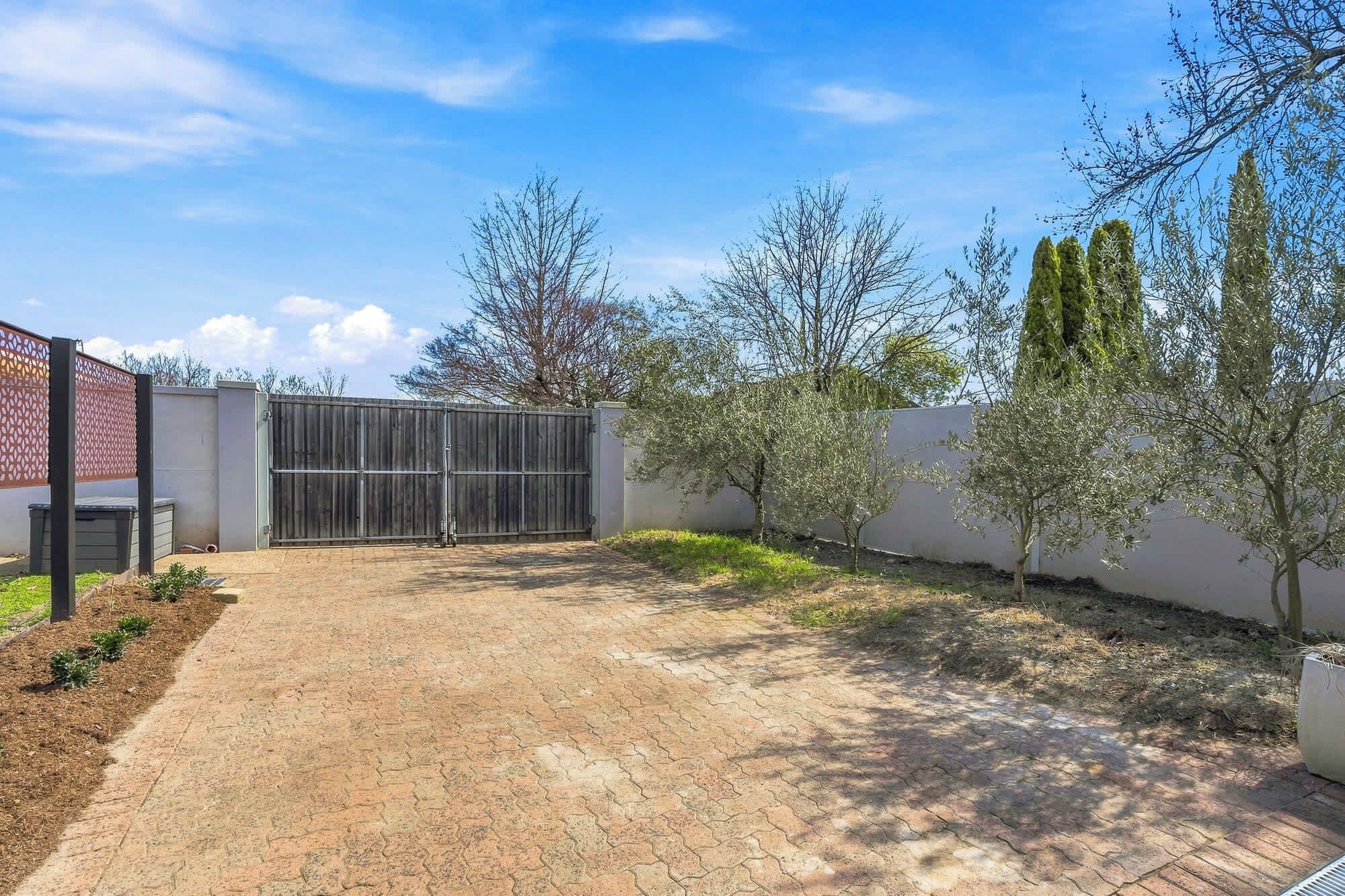 A paved courtyard with a metal gate, surrounded by trees and a white wall.