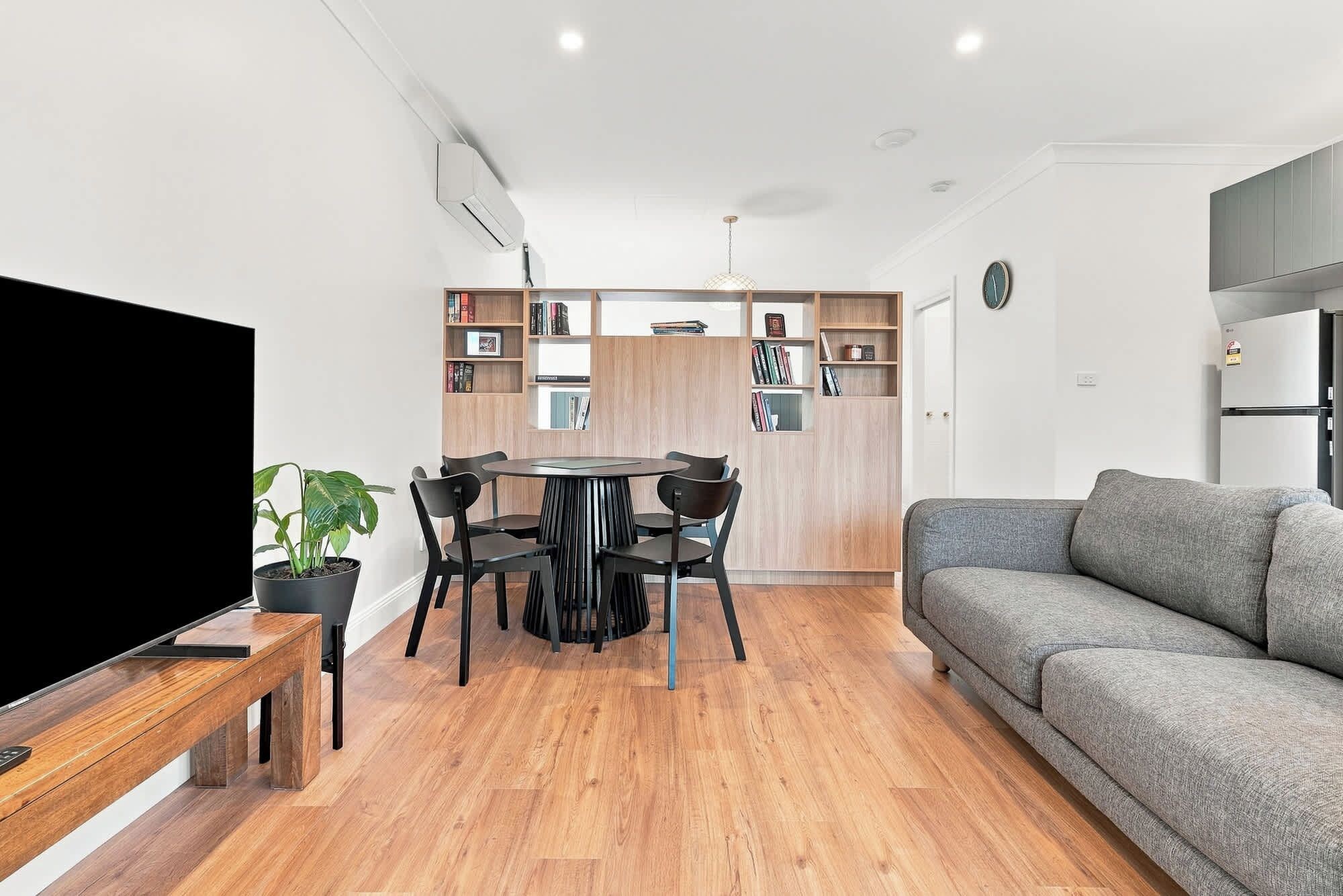 Ein modernes Wohnzimmer mit einem grauen Sofa, einem Holzesstisch mit Stühlen, einem Fernseher auf einem Schrank und einem Bücherregal.