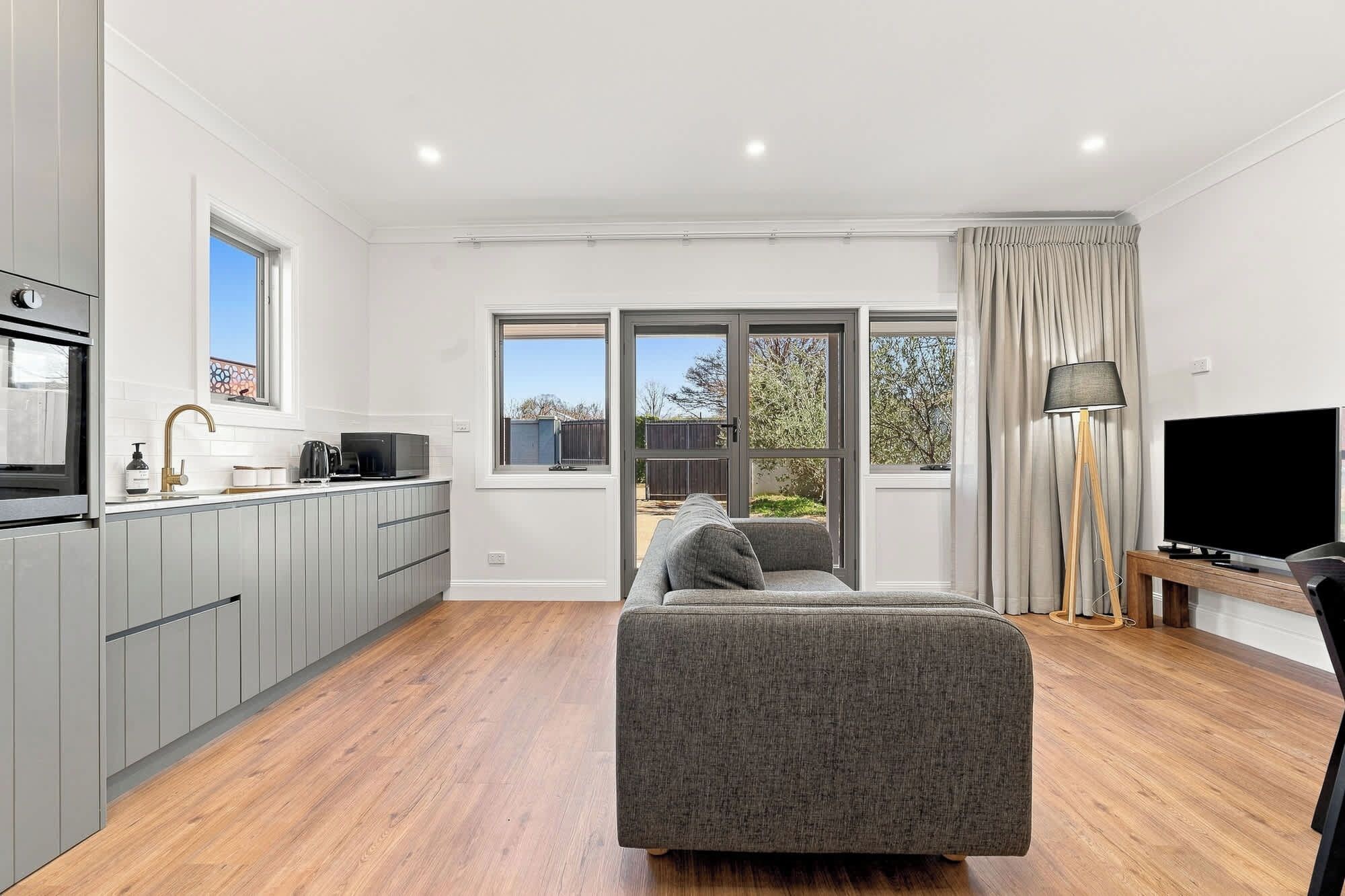 A modern living space with a kitchen, a grey sofa, a TV, and a large window with curtains.