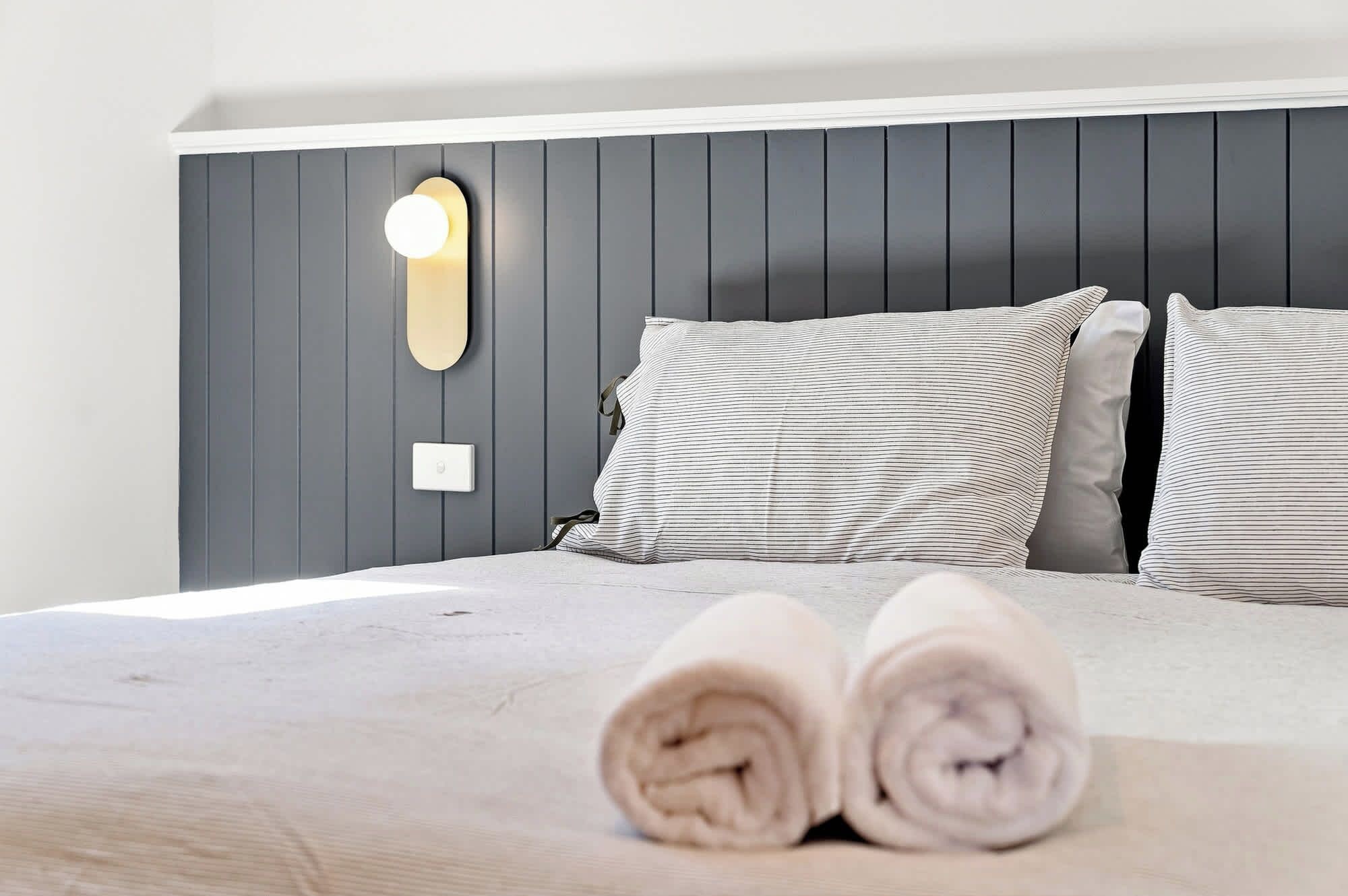 Cama bem arrumada com toalhas dobradas, luminária na parede e almofadas listradas.