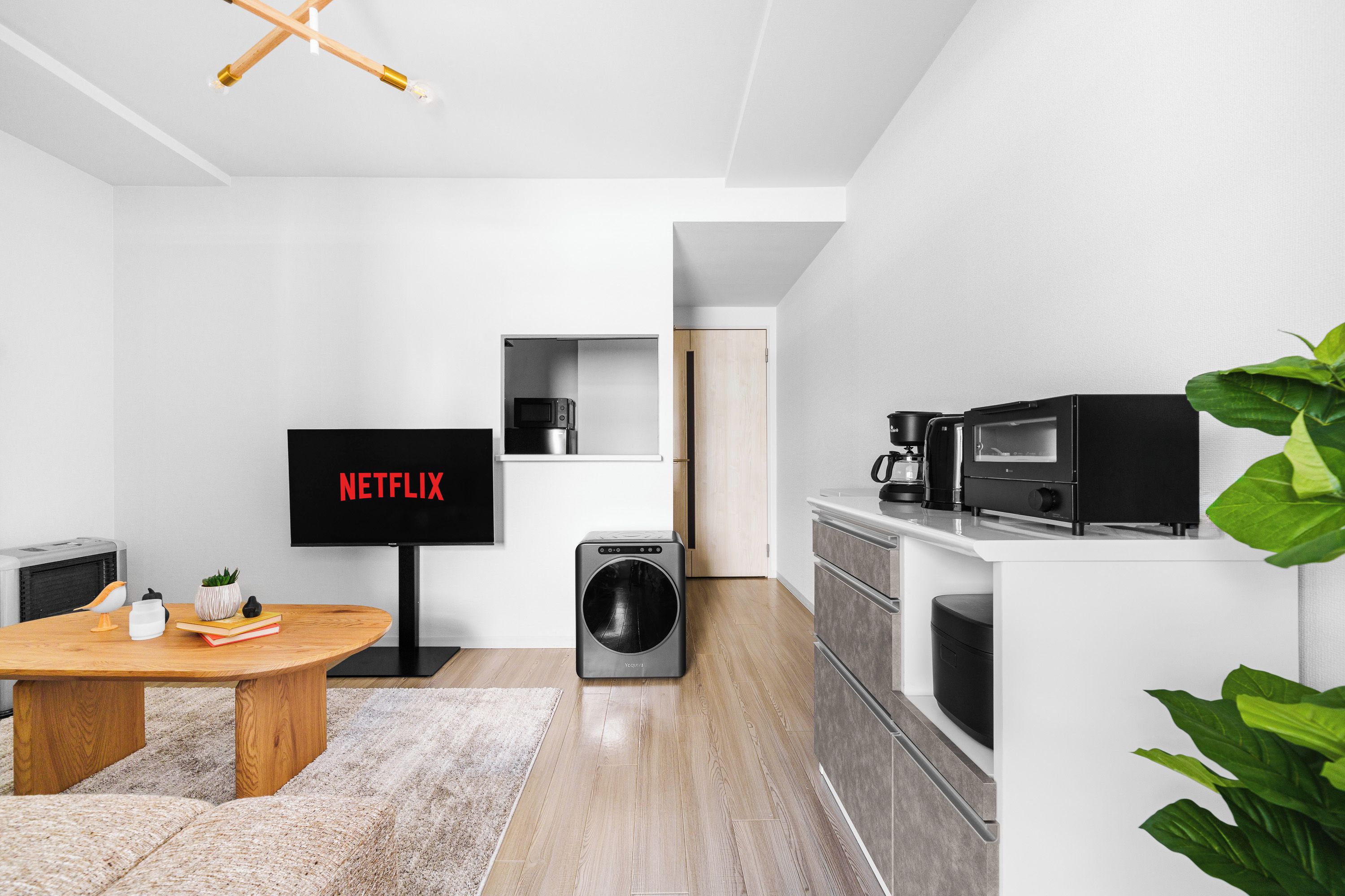 A modern living room with a flat-screen TV, a washing machine, and a kitchen area with appliances.