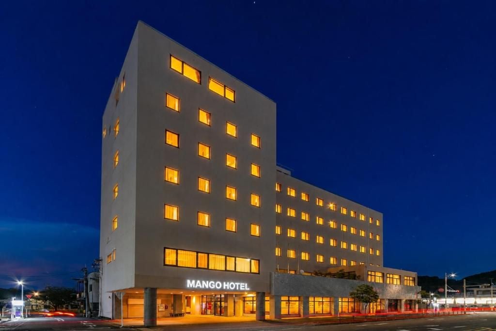 Un hôtel à plusieurs étages, doté d’une façade sobre, de fenêtres illuminées et d’une enseigne indiquant « MANGO HOTEL » à l’entrée.