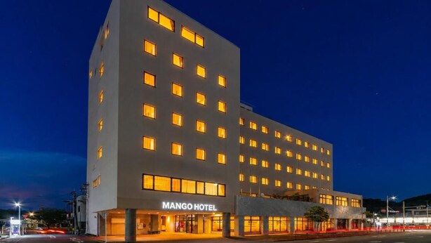 Monikerroksinen hotelli, jossa on tasainen julkisivu, valaistut ikkunat ja sisäänkäynnin yläpuolella oleva kyltti, jossa lukee ”MANGO HOTEL”.