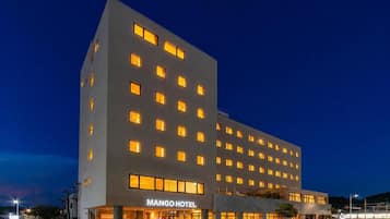 Un hôtel à plusieurs étages, doté d’une façade sobre, de fenêtres illuminées et d’une enseigne indiquant « MANGO HOTEL » à l’entrée.