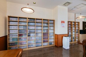 A library interior with multiple bookshelves, a wooden table, and a white air purifier. - Mango Hotel Nichinan (Nichinan)