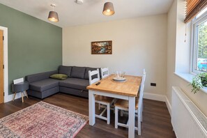 Una sala moderna con un sofá gris, una mesa de comedor de madera con sillas, un cuadro en la pared y una ventana con persianas.
