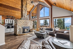 A spacious living room with a stone fireplace, leather sofas, and large windows offering a view of the mountains. - Big White is Calling - Full House with Hot Tub (Kootenay Boundary)