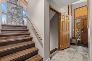 Un escalier en bois mène à un mur de pierre, une porte en bois, et on peut apercevoir un autre mur de pierre à l’extérieur.