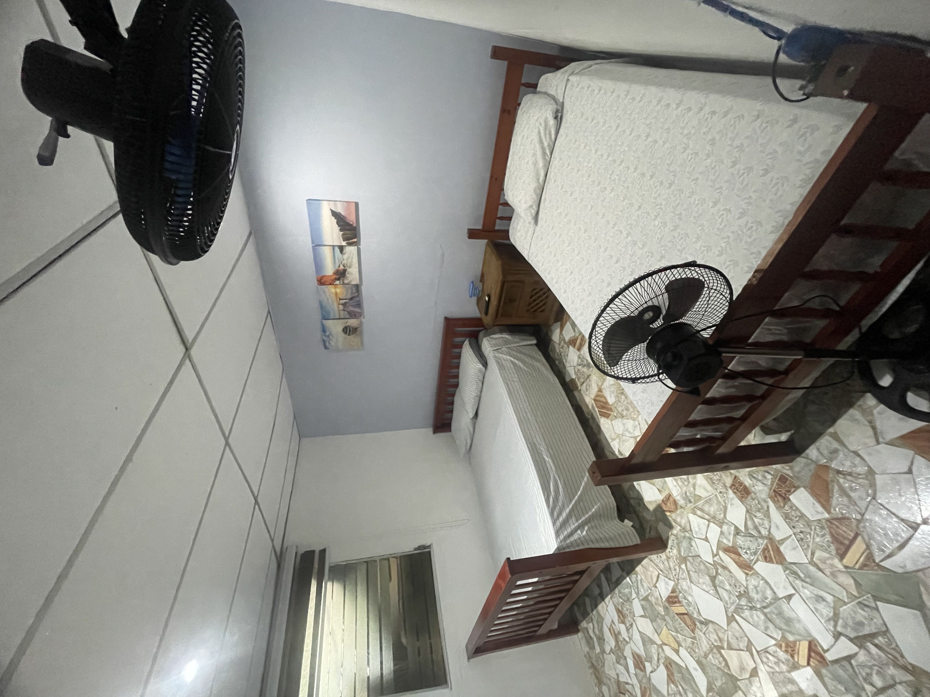 Quarto com cama, ventoinha, ar condicionado na parede e uma pequena janela.