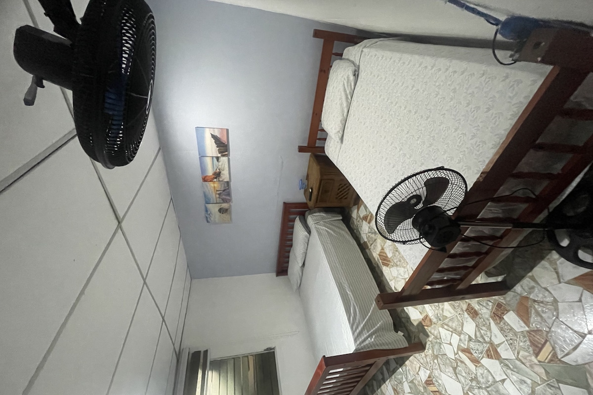 A room with a bed, a standing fan, a wall-mounted air conditioner, and a small window.