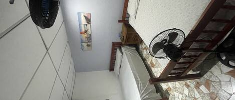 Una habitación con una cama, un ventilador de pie, aire acondicionado instalado en la pared y una ventana pequeña.