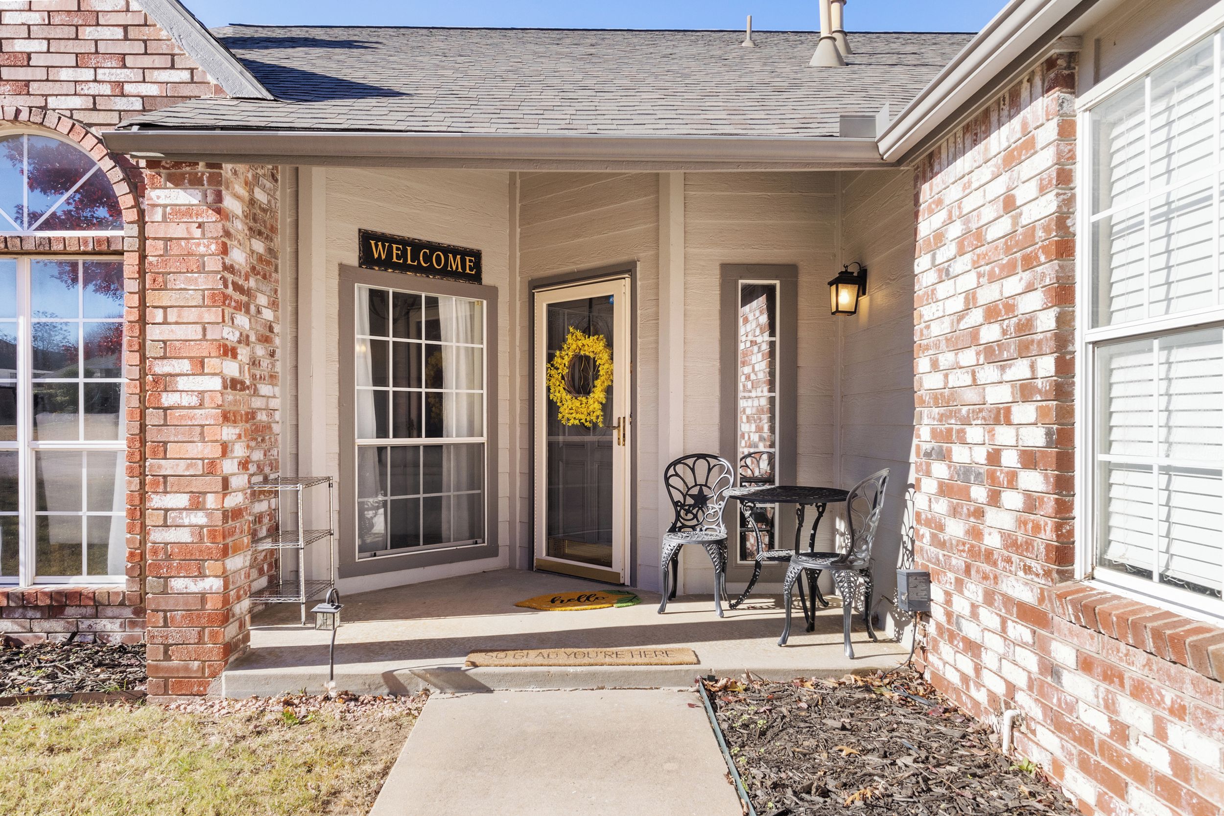 A brick house with a front porch, a 'WELCOME' sign, a wreath, and outdoor furniture.