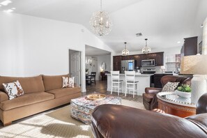A modern living room with a brown sofa, patterned ottoman, and leather armchair. A kitchen with dark wood cabinets and white appliances is visible in the background. - Furnished Executive Stay Offers 2 Living, 2 King 1 Queen Private Primary Suite (Tulsa)