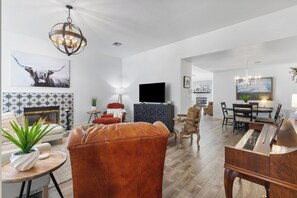 A spacious living room with a fireplace, a large TV, a piano, and a dining area with a chandelier. - Furnished Executive Stay Offers 2 Living, 2 King 1 Queen Private Primary Suite (Tulsa)