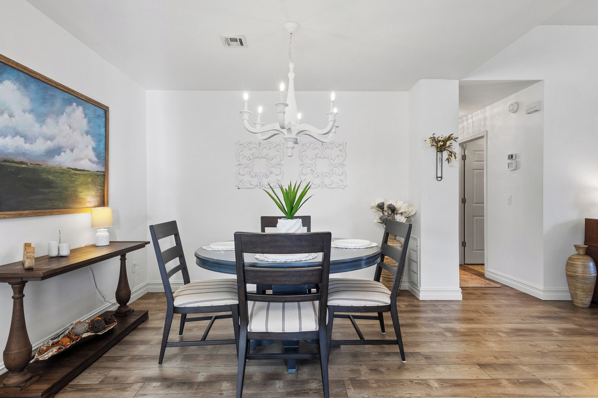 A dining area with a round table, four chairs, a chandelier, a painting, and a vase with flowers.