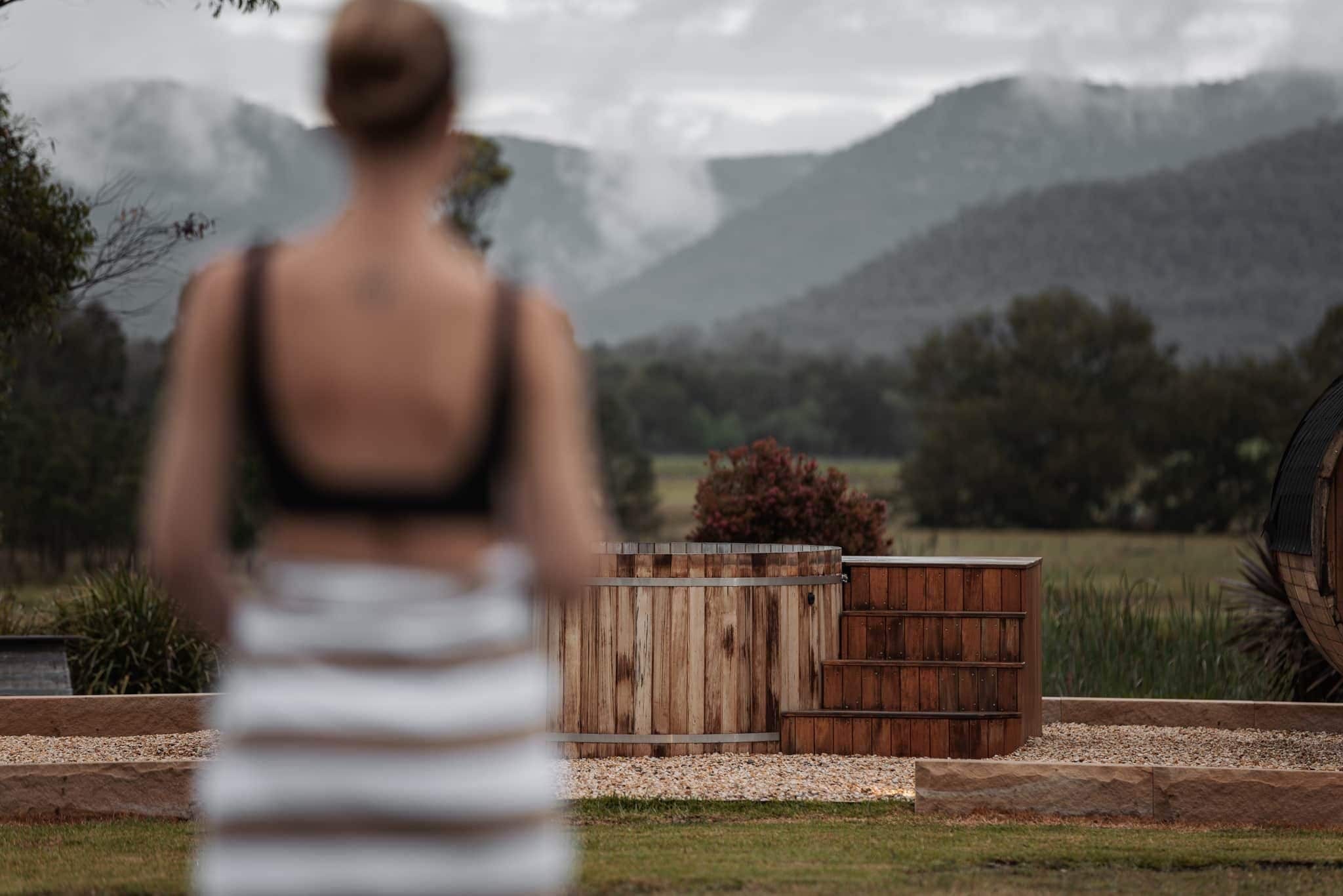 Une personne se tient dans un jardin, près d’une structure en bois, avec une vue sur les montagnes.
