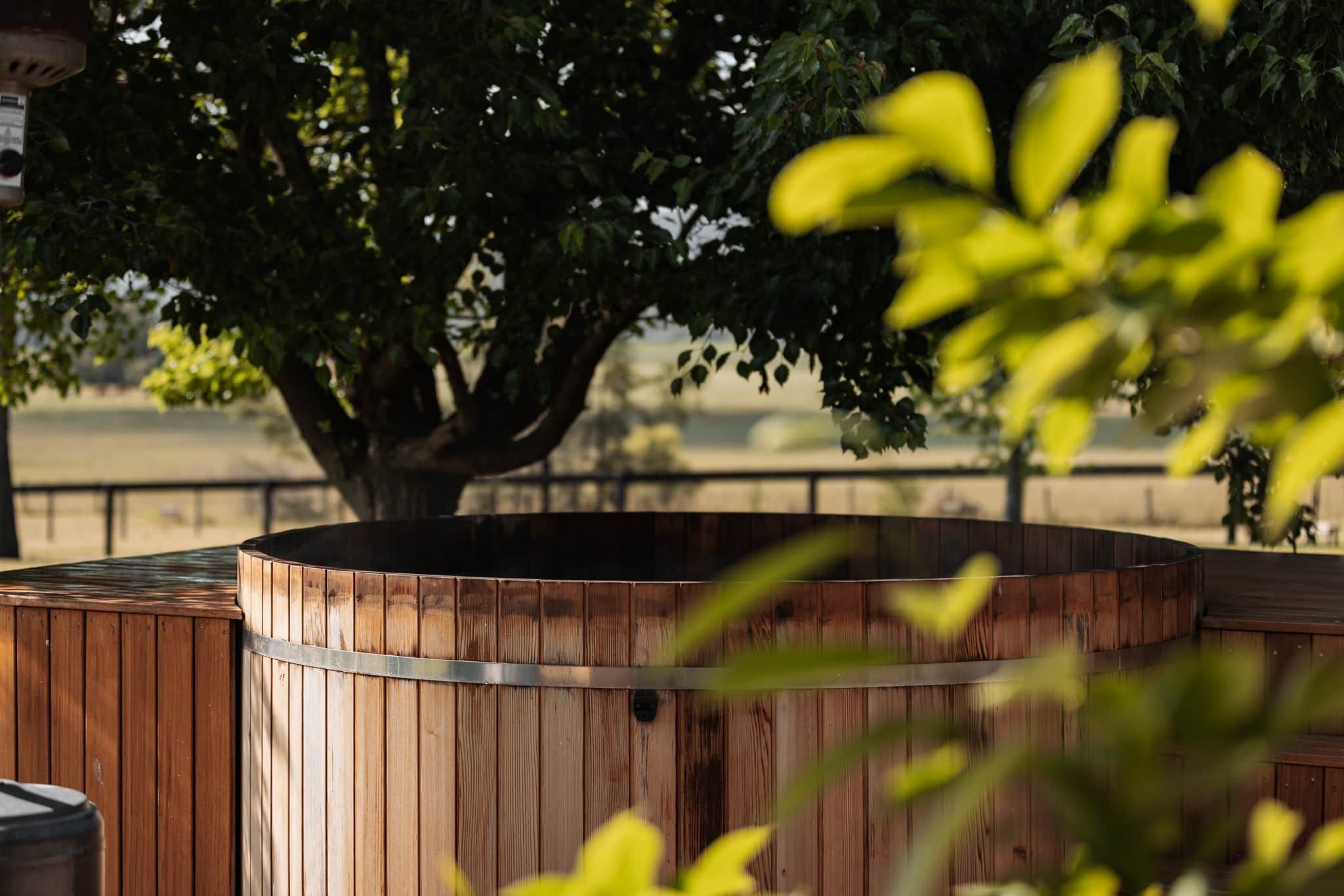 A wooden barrel structure, possibly a hot tub, with a metal band around it, set against a backdrop of a tree and a fence.