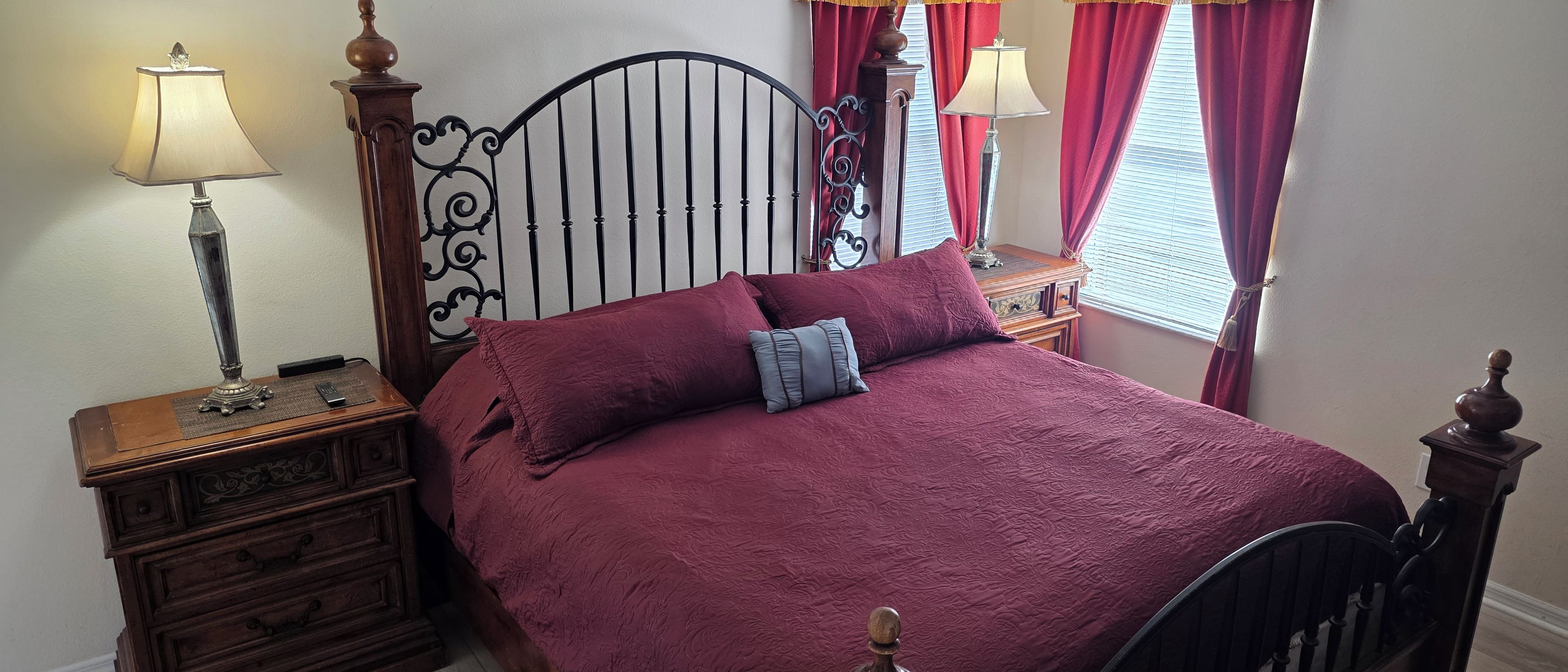 A bedroom with a four-poster bed, two bedside tables with lamps, a dresser, and a window with red curtains.