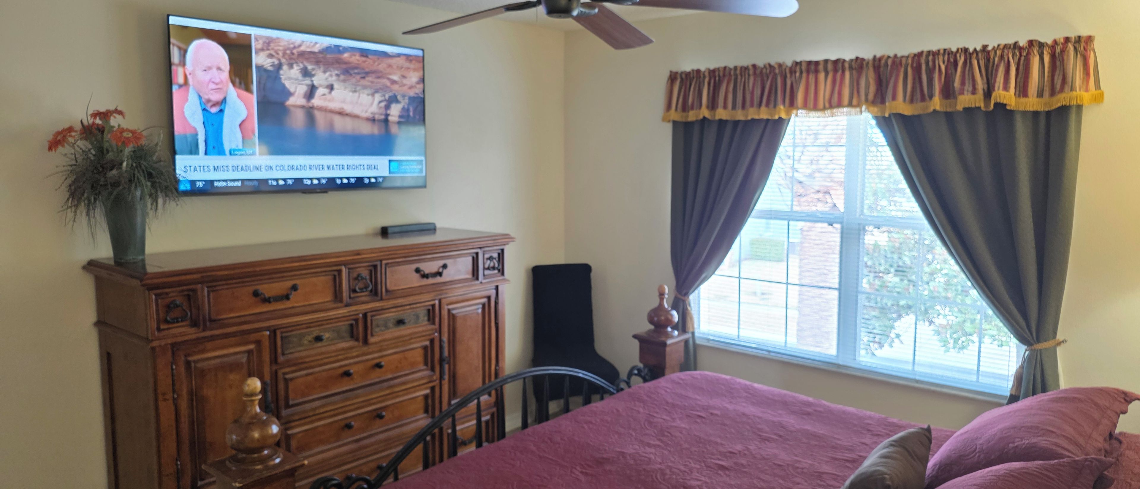 A bedroom with a bed, a dresser, a television mounted on the wall, and a window with curtains.
