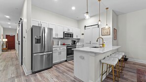 A modern kitchen with stainless steel appliances, a central island with bar stools, and pendant lighting. - Crimson Cajun | Where Southern Comfort Meets Football (Tuscaloosa)