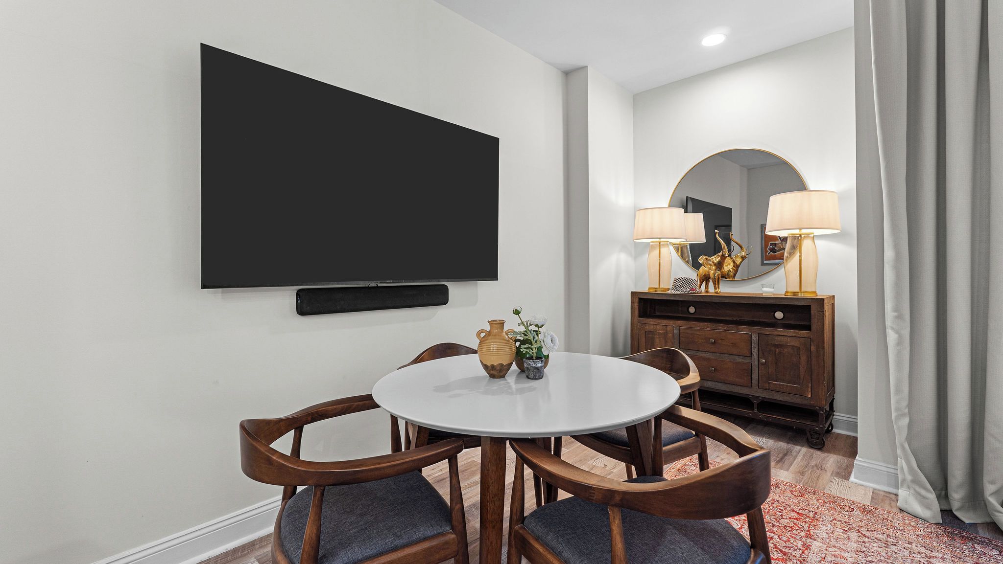 Habitación de hotel con televisión de pantalla plana montada en la pared, una mesa redonda con sillas, un armario de madera con lámparas y una alfombra estampada.