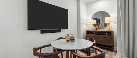 Habitación de hotel con televisión de pantalla plana montada en la pared, una mesa redonda con sillas, un armario de madera con lámparas y una alfombra estampada.