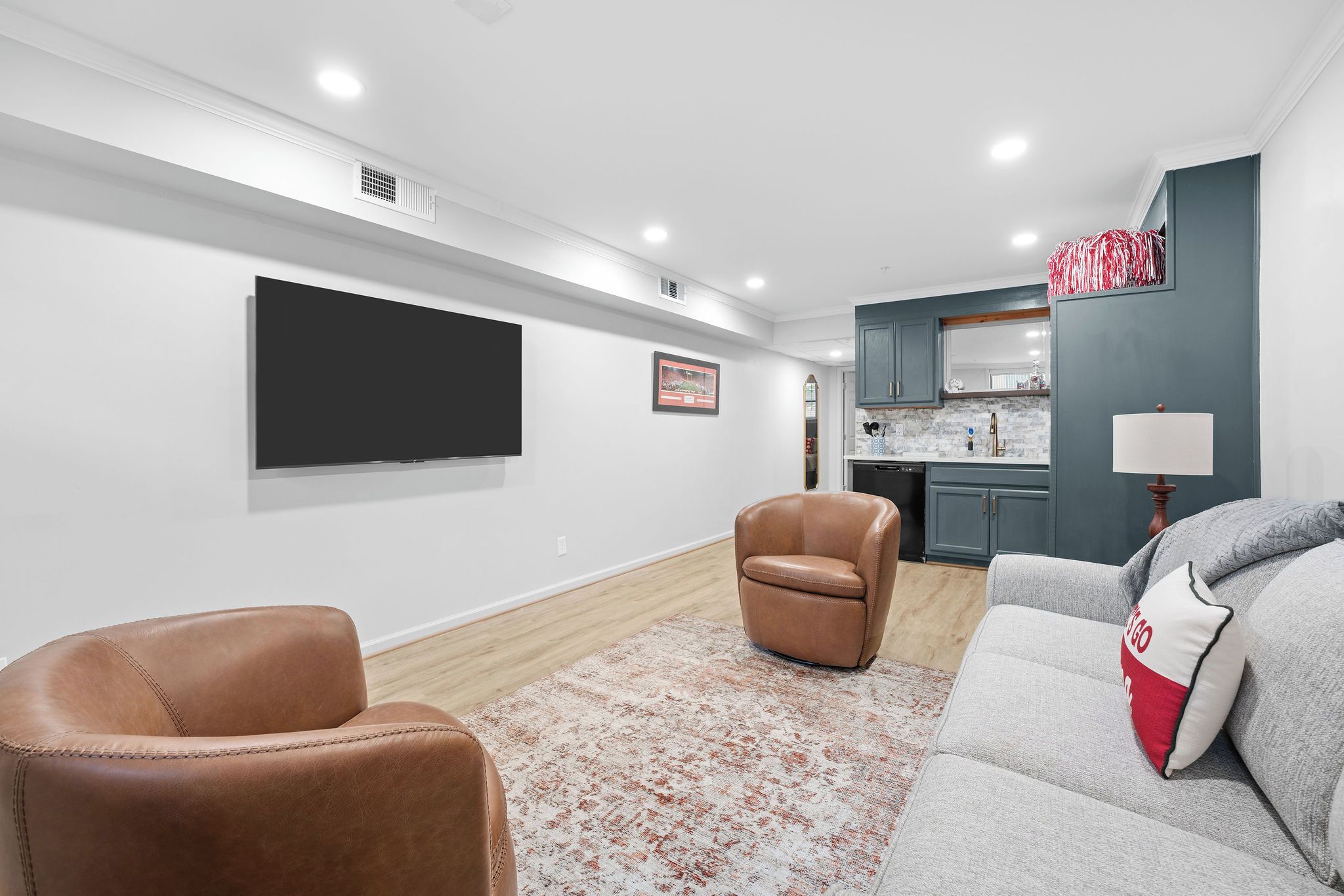 A modern living room with a flat-screen TV, leather armchairs, a grey sofa, and a kitchen in the background.