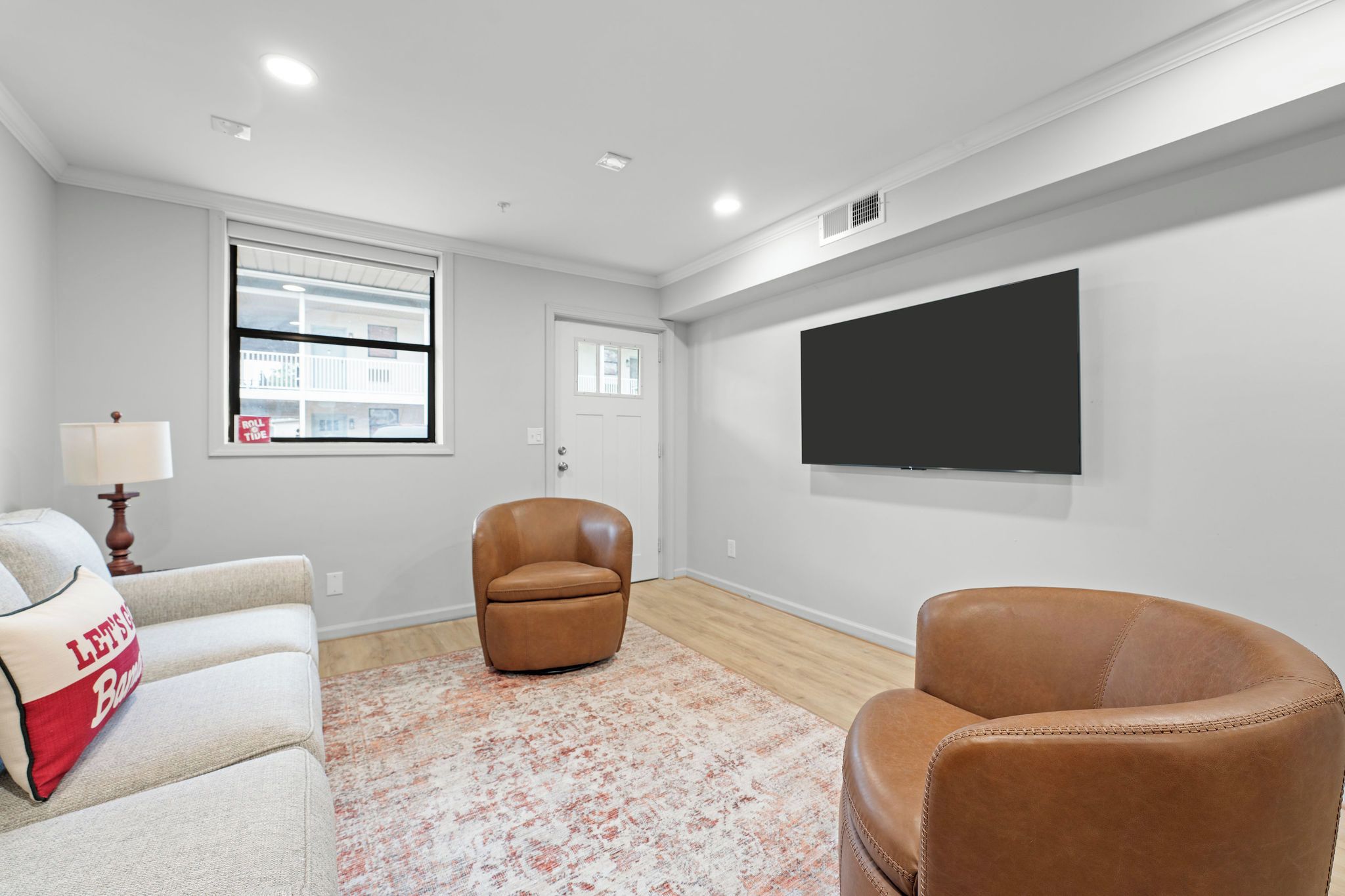 A living room with a sofa, two leather chairs, a TV mounted on the wall, and a window with a view of buildings.