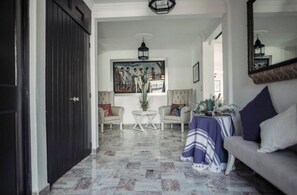 Un couloir avec une porte sombre, deux fauteuils, une petite table ronde avec une plante et un canapé recouvert d’un plaid à rayures.