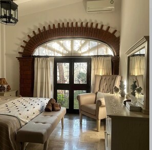 Une chambre avec une grande fenêtre en arc de cercle, un lit recouvert d’une couvre-lit à motifs, une chaise et une commode.