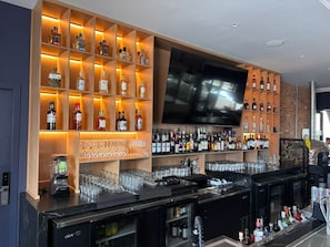 Un espace bar avec des étagères remplies de bouteilles d’alcool, un téléviseur et un comptoir avec des verres et un mixeur.