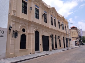 A building with multiple windows and doors, some with shutters, and a street in front.
