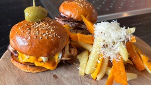 Un plato con dos hamburguesas de queso y pepinillos, acompañado de papas fritas y queso rallado.