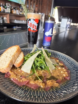 Une assiette de nourriture accompagnée de pain et de légumes verts, un verre d’une boisson sombre et une canette de boisson, le tout dans un cadre de bar.