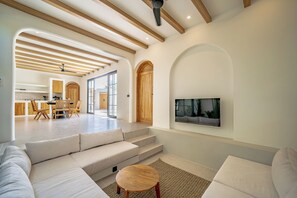A spacious living area with a large sofa, a coffee table, and a TV mounted on the wall. There is a staircase leading to an upper level, and a dining area with wooden furniture is visible in the background. - Palmera Moana Uluwatu (Pecatu)