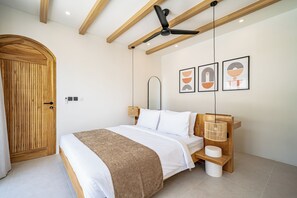 A bedroom with a bed, wooden headboard, ceiling fan, wall-mounted light, and artwork. - Palmera Moana Uluwatu (Pecatu)