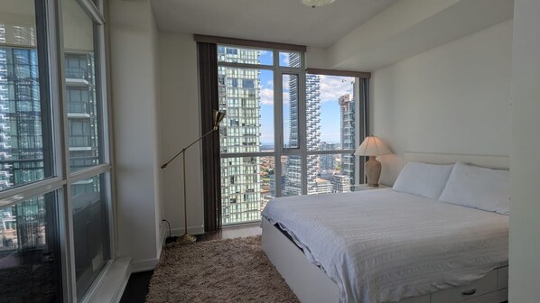 A bedroom with a large window, a bed, a lamp, and a view of high-rise buildings.