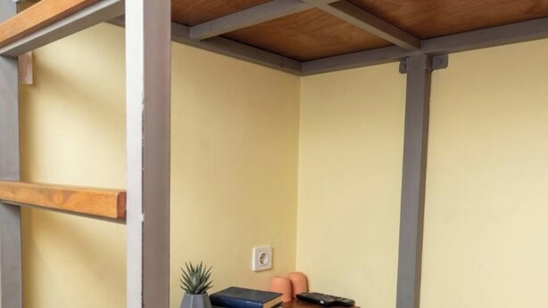 Una habitación con techo de madera, pared amarilla, una planta pequeña, un libro y un control remoto sobre una mesa.