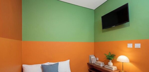 Una habitació amb combinació de colors verd i taronja a les parets, un televisor de pantalla plana penjat a la paret, una taula de nit amb una làmpada i un llit amb almoadins.