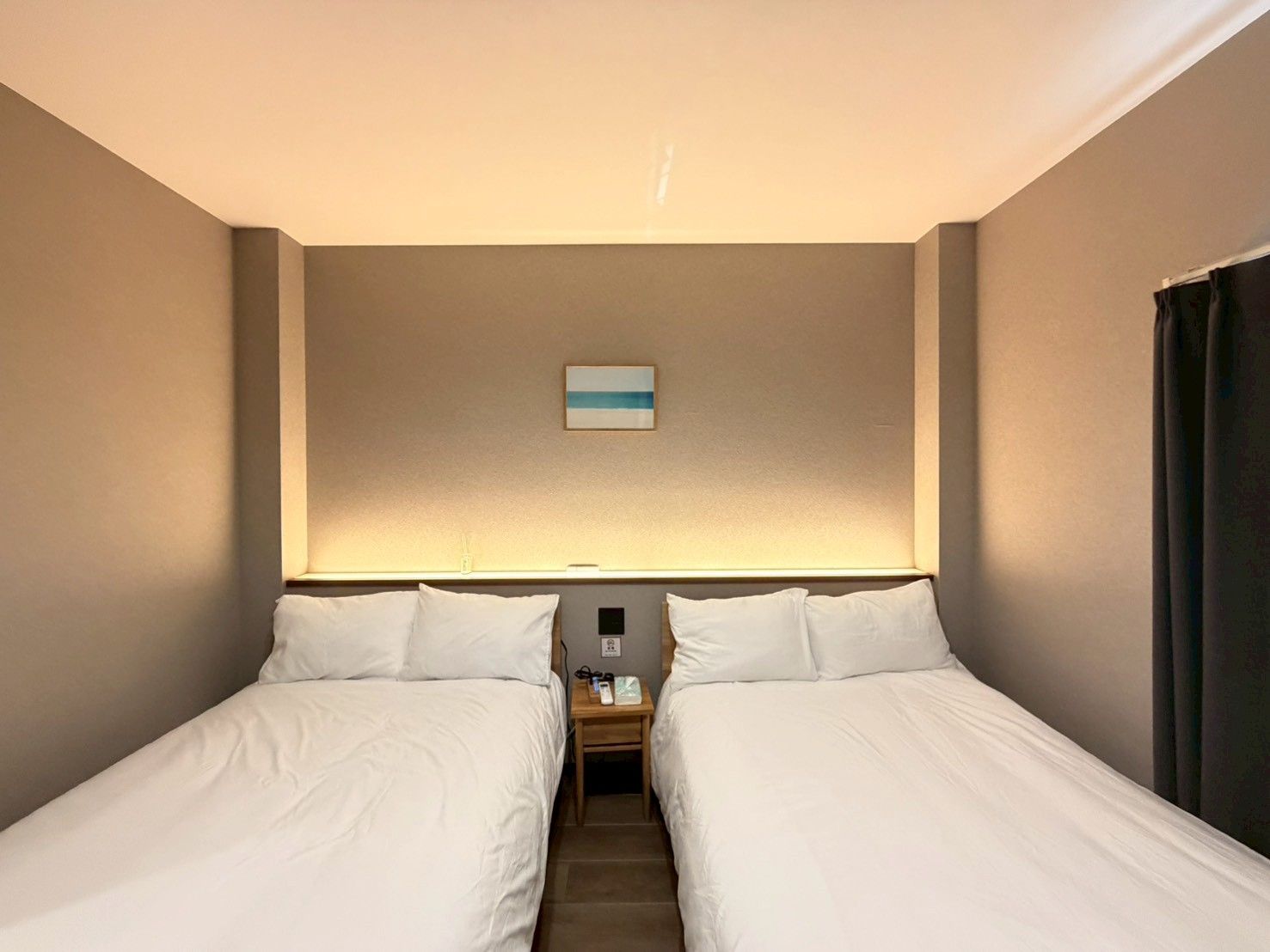A hotel room with two beds, a nightstand, a wall-mounted lamp, and a small painting.