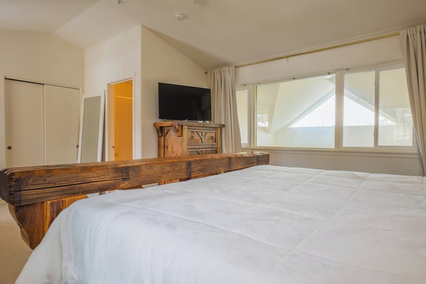 Une chambre à coucher avec un grand lit, une tête de lit en bois, un téléviseur posé sur un meuble en bois et une fenêtre avec des rideaux.