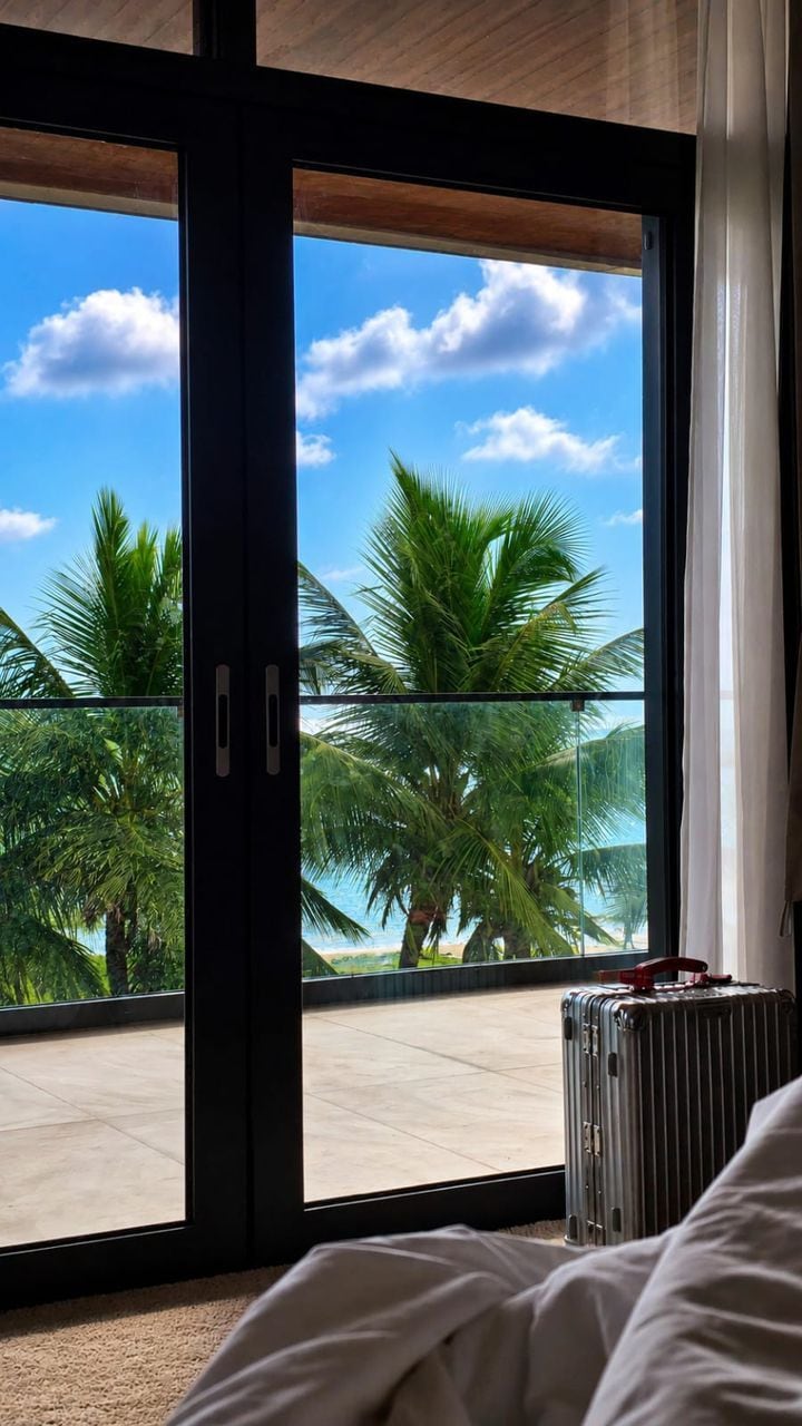 A room with a large sliding glass door leading to a balcony with a view of palm trees and a beach.