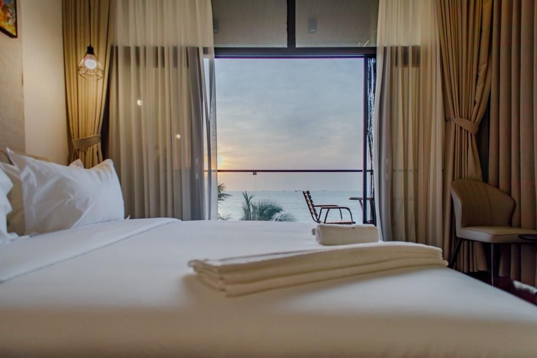 A hotel room with a bed, a chair, a small table, and a view of the sea through a large window.