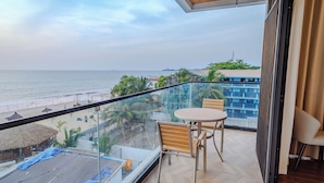Een balkon met glazen hekwerk, uitzicht op een strand, een tafel met stoelen, en een beeld van de zee en verre gebouwen.