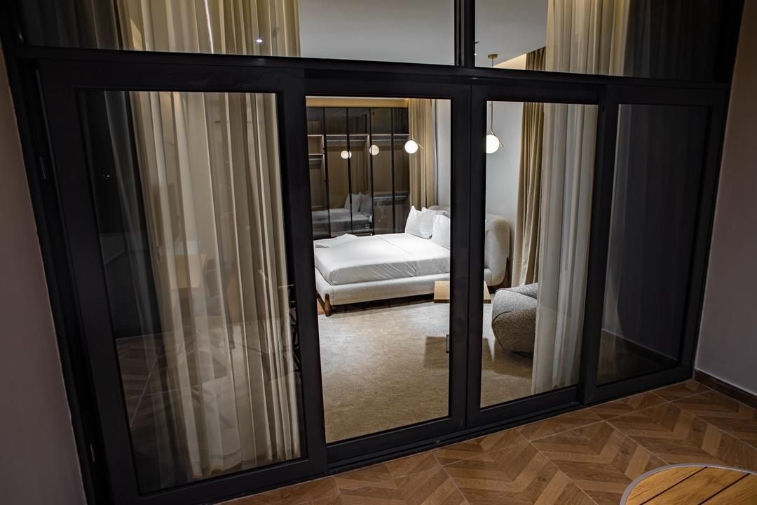 Una habitación de hotel moderna con un amplio ventanal corredero que da a un balcón, una cama con ropa de cama blanca y una zona de estar con una silla.