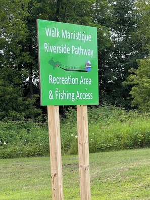 A green signboard for the Walk Manistique Riverside Pathway, indicating it is a recreation area with fishing access.