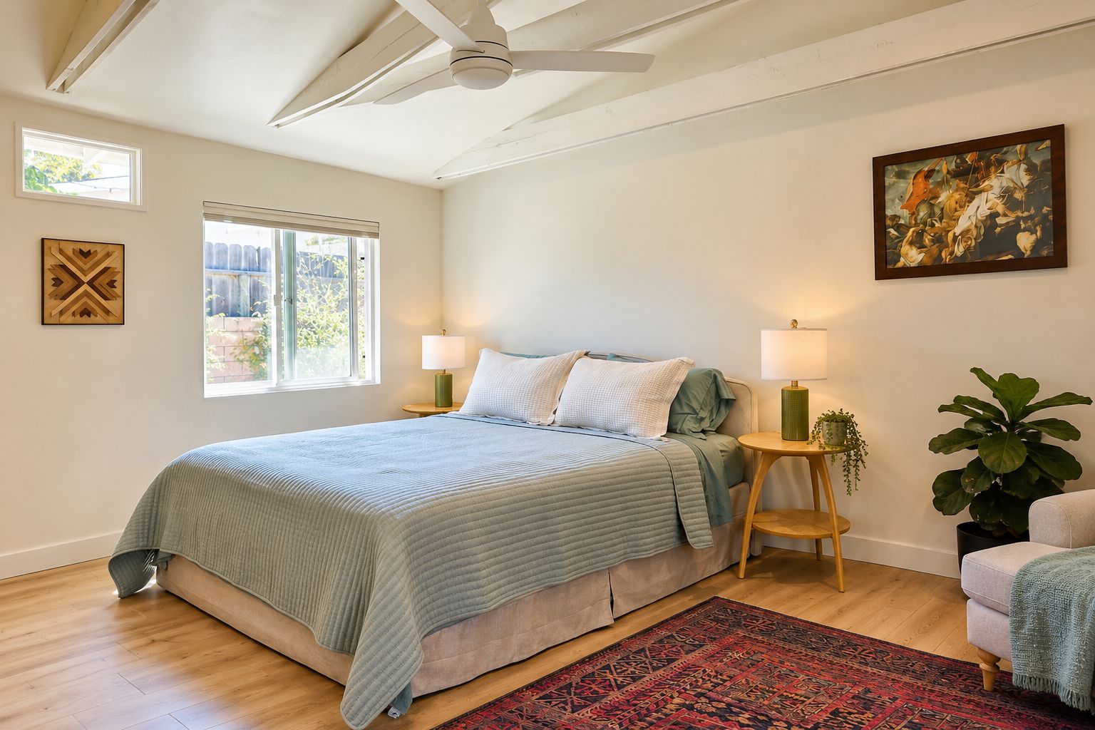 A bedroom with a bed, two bedside lamps, a window, a ceiling fan, a painting, and a potted plant.