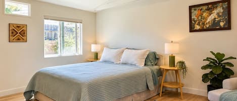 A bedroom with a bed, two bedside lamps, a window, a ceiling fan, a painting, and a potted plant.