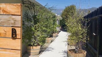 Un sendero bordeado por estructuras de madera y plantas en macetas, que lleva a un edificio con una puerta marcada con una letra.