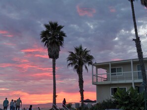 Un immeuble situé en bord de mer, doté d’un balcon offrant une vue sur l’océan, les palmiers et le ciel au coucher du soleil.